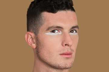 Fototapeta premium Closeup portrait of a strong young White man with short brown hair, light makeup and white eyeshadow sitting by himself inside a studio with a pecan background wearing a black shirt.