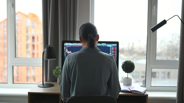 Back view of multiracial woman freelancer sitting at the table and typing on the keyboard, using trendy computer for remote work. Modern home office concept