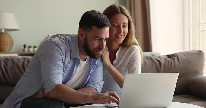 Smiling young male female family couple relax hug on comfy couch before laptop screen choose interior design for new home online. Happy bonding husband wife use wifi to watch funny video content on pc