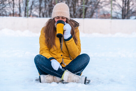 Young woman in ice skates sitting on the snow and drinking coffee. 