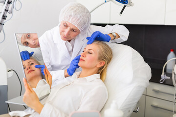 Smiling adult woman looking at mirror while elderly female cosmetician showing results of hardware facial procedure in medical esthetic office