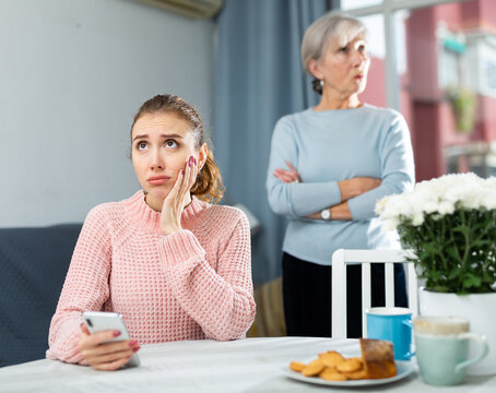 Offended young woman sitting separately at table at home having problems in relationship with aged mother.