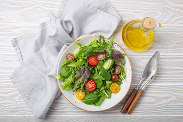 Vegetables healthy salad with red yellow cherry tomatoes and green salad leafs on white rustic ceramic plate, olive oil bottle on white wooden rustic background top view, healthy food and diet concept
