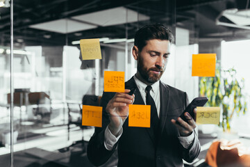 Successful businessman rewrites a task on a glass board with colored stickers, from phone and tablet