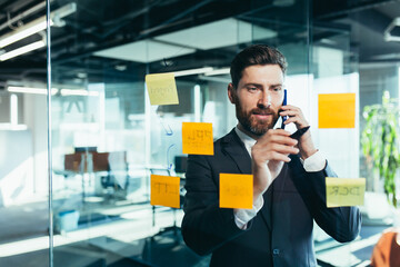 businessman in reflection at a glass task board, a man talking on the phone, hanging plans on...