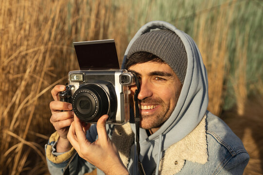 Close-up young handsome man smiling taking pictures of holidays with instant camera wearing a beanie and blue jacket