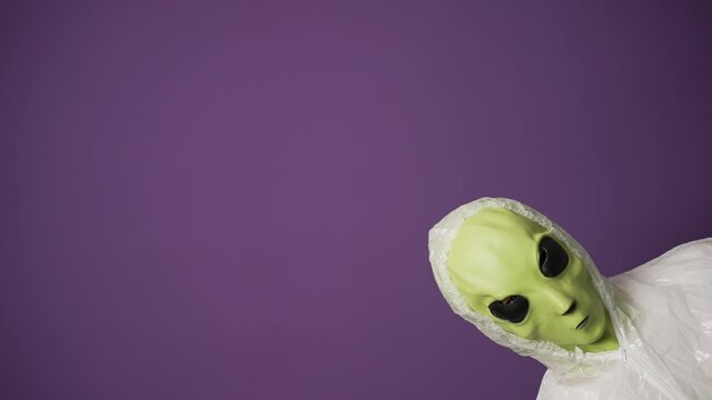 A funny green alien in a white suit peeks out of the corner of the frame against a purple background. Alien invasion