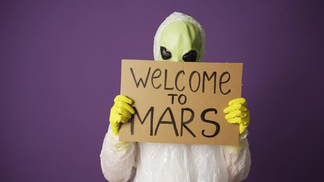 Green positive alien in white suit raises his toddler with the words welcome to mars on purple background