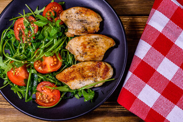 Roasted chicken breasts and salad with arugula and cherry tomatoes in a black plate. Top view