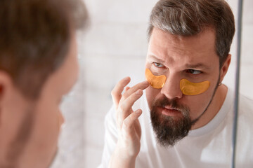 Portrait of man in white tshirt applying eye patches on his face at bathroom. Self care morning procedure