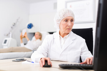 Portrait of professional experienced female cosmetologist working with laptop in modern aesthetic medicine office, writing patient examination results