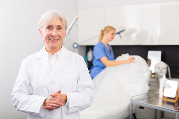 Senior woman cosmetologist standing in her office, looking at camera and smiling. Her assistant nurse working in background.