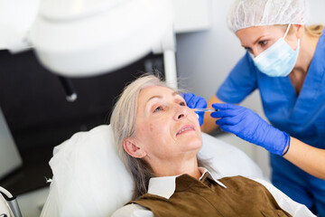 Portrait of elderly female client of aesthetic cosmetology clinic during procedure of face mesotherapy. Anti aging skin care concept