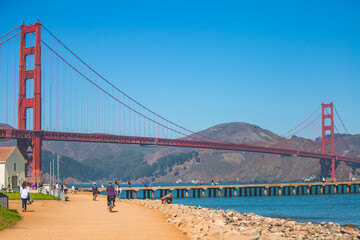 People walking on San Francisco Bay Trail next to Warming Hut Bookstore, Torpedo Wharf, and Golden Gate Bridge on a clear sunny day.