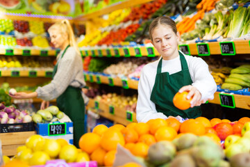 Focused young girl working at family fruit and vegetable shop, arranging ripe oranges on showcase..