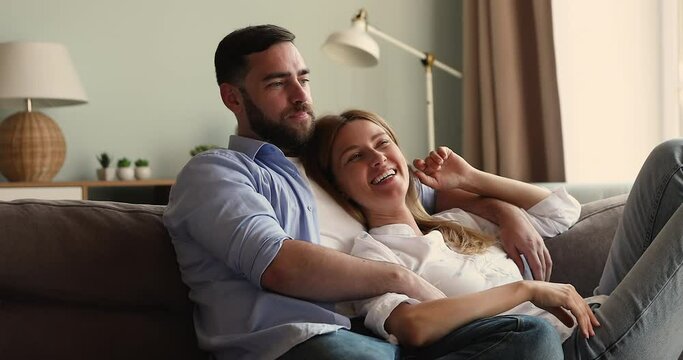 Bonding millennial husband wife cuddle on comfy couch engaged in warm conversation create plans for future. Affectionate inspired young family couple share ideas dreams resting on sofa at living room