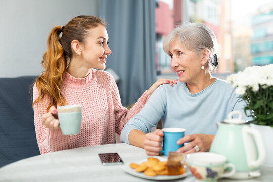 Happy young girl and elderly woman sitting at home table, drinking tea and talking. Friendly conversation between mother and daughter.