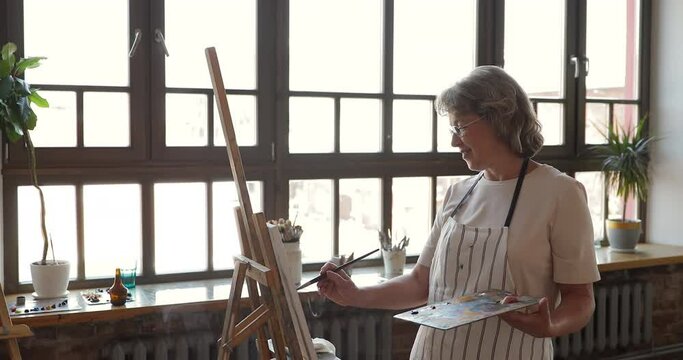 Smiling mature adult female experienced painter engaged in creative process of drawing picture add more details with expressive brushstrokes. Confident aged woman artist paint in oils at comfy studio