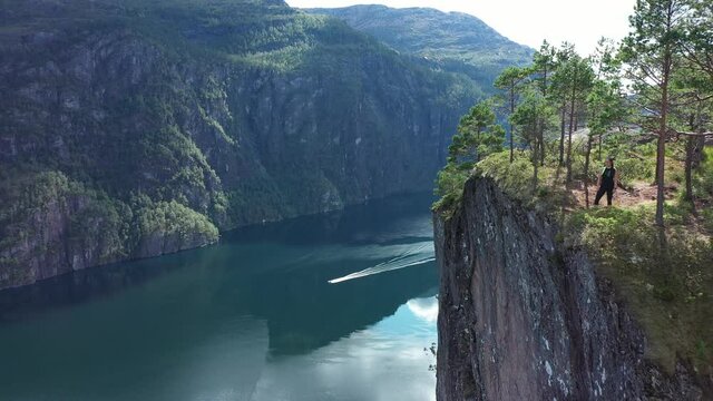 Woman enjoying spectacular view from edge of tall mountain cliff Slottet in Modalen Norway - Forward moving aerial close to cliff in stunning fjord landscape with boat passing on fjord below - Norway