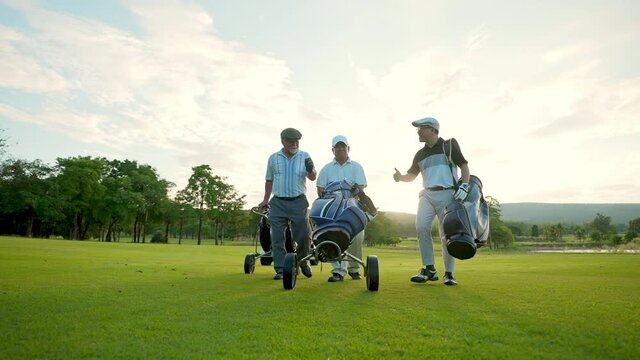 Group of Asian people businessman and senior CEO enjoy outdoor sport golfing together at country club. Healthy men golfer holding golf bag walking on fairway with talking together on summer vacation