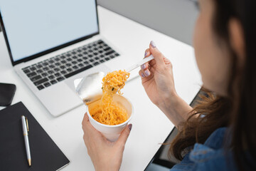 Freelance Asian businesswoman eating instant noodles while working on laptop at home