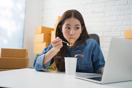 Young asian female at laptop computer and eats instant ramen noodles late in evening.