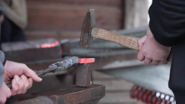 Two caucasian male blacksmiths forging and hammering on a piece of red hot metal at a smithy on an anvil