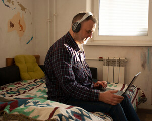 A Man of European appearance in large laptop headphones on his lap a plaid shirt sitting on a sofa at home. Remote work. On-line courses.