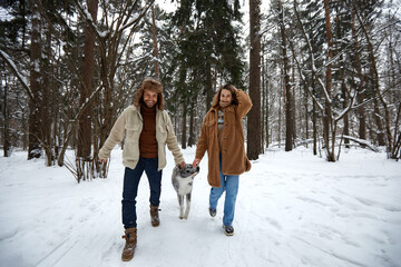 Happy young couple in winterwear playing with purebred siberian husky and having fun in snowbound pine forest