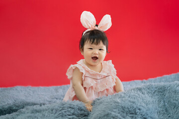 Fototapeta premium Asian Baby in Easter bunny hairband. Asian happy baby smiling and sitting on red background. Cute 6 months baby start sitting with copy space as Easter concept, baby or kid department in hospital.