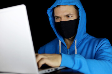 Man in mask and blue hoodie sitting with laptop on black background. Concept of cyber crime, hacking and technology