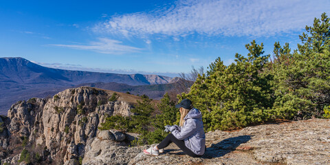 A girl in a gray jacket and a black cap near green pine trees on the top of a mountain against a blue sky in the clouds looks into the distance at a beautiful valley