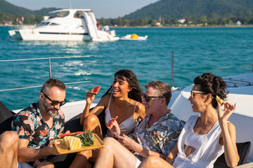 Group of man and woman friends enjoy outdoor party eating fresh fruit together while catamaran boat...