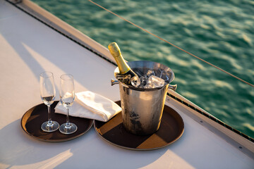 Champagne bottle in ice bucket with champagne glass for serving to passenger tourist on luxury catamaran boat sailing in the ocean at summer sunset. Tropical travel vacation sail yacht trip concept - obrazy, fototapety, plakaty