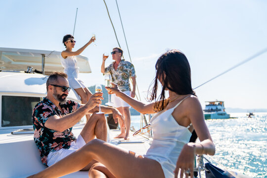 Group of Caucasian people friends enjoy luxury party drinking champagne together while catamaran boat sailing in the ocean. Man and woman relax outdoor lifestyle sail yacht on tropical travel vacation