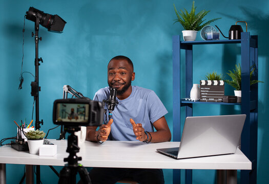 Smiling vlogger talking with audience in front of recording digital video camera during online live show. Content creator interacting with fanbase in studio looking at dslr live video podcast setup.