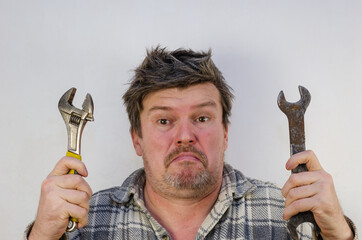 An adult Man with a perplexed face holds two wrenches.