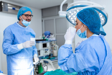 Naklejka na ściany i meble Team of surgeons with surgical mask at operating room drinking water after successful operation . Young woman doctor in surgical uniform in hospital operation theater.