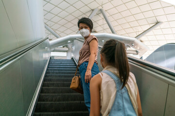 Asian woman wearing face mask and talking with her daughter while using escalator in subway station during coronavirus or covid-19 outbreak. social distancing, quarantine or new normal concept