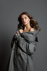 Beautiful woman in a beige bodysuit and a long gray raincoat poses standing on a gray background. Long beautiful legs