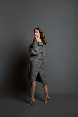 Fototapeta premium Beautiful woman in a beige bodysuit and a long gray raincoat poses standing on a gray background. Long beautiful legs
