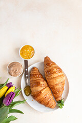 Breakfast with fresh croissants, chocolate cream, jam and colorful tulips on light stone background. Beautiful romantic composition. Top view.