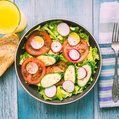 Vegan salad with radishes, tomatoes, cucumber and lettuce in a plate on a table with toast and citrus juice, top view