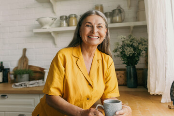Cheerful smiling mature female in good mood drinking tea on kitchen. Joyful senior lady in yellow...