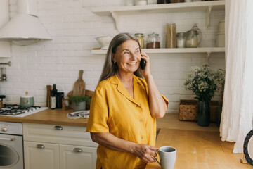 Beautiful grandmother in casual yellow clothes talking to her son on smartphone, looking aside, standing half-turned to camera near table counter in bright kitchen, having rest, spending leisure time - 479510054