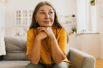 Mature woman sitting on couch putting chin on crossed hands, looking aside with positively...