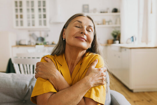 Horizontal indoor portrait of happy elderly woman hugging herself, giving support, showing self-respect and love, sitting on couch with closed eyes in yellow shirt. Human emotions, body language