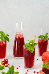 Refreshing pomegranate juice in glasses with mint and glass straw, pitcher of juice and ice, cold drink