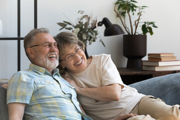 Fototapeta premium Happy affectionate 60s mature family couple looking in distance, resting on comfortable sofa, enjoying communicating, dreaming planning future or watching funny TV shows together on weekend at home.