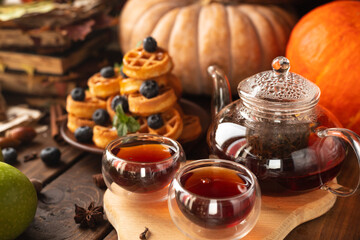 many waffles are stacked on a plate, decorated with mint leaves and blueberries. Tea in a glass teapot and two small bowls